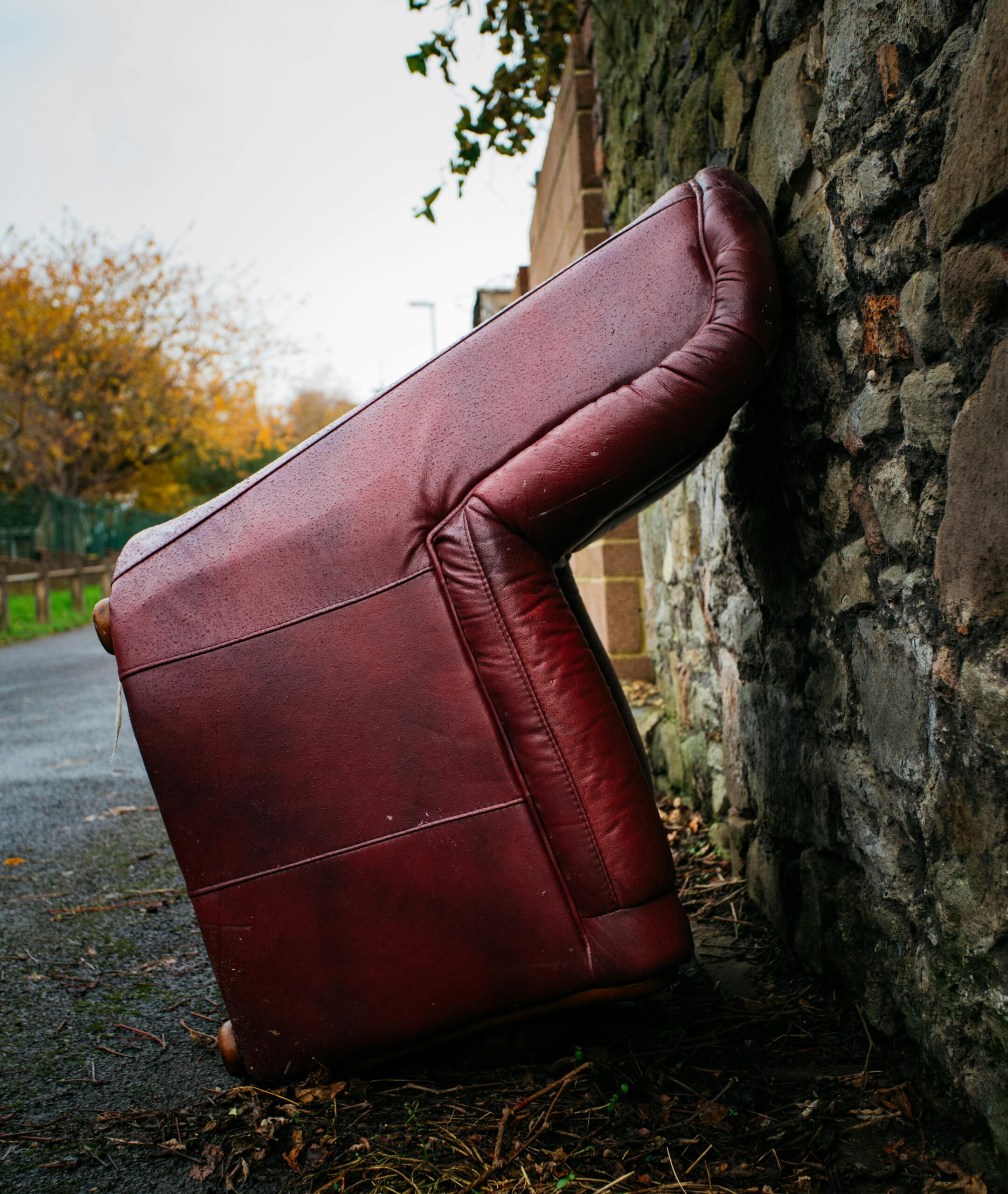 An abandoned red leather sofa leaning against a stone wall in an outdoor setting.